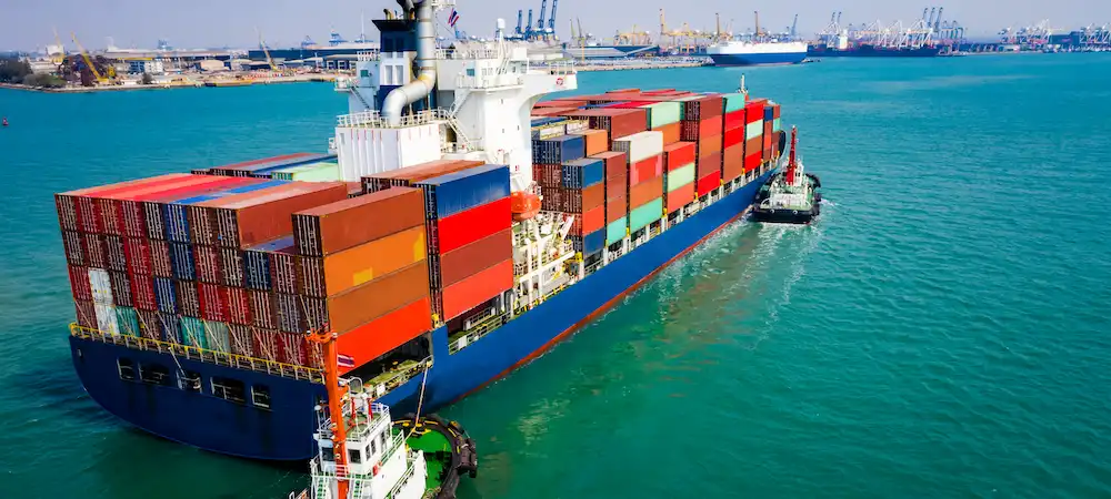 A large cargo container ship loaded with colorful shipping containers being guided by a tugboat in a busy port, representing international logistics and expedited shipping methods.