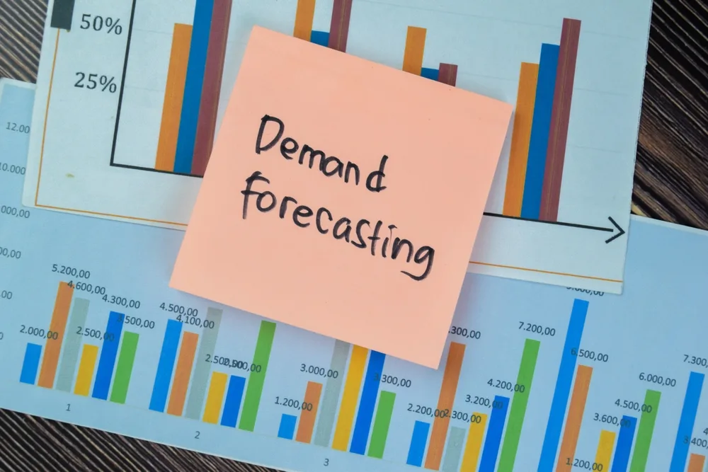 Sticky note with handwritten "Demand forecasting" text placed on top of financial supply chain data charts and bar graphs.