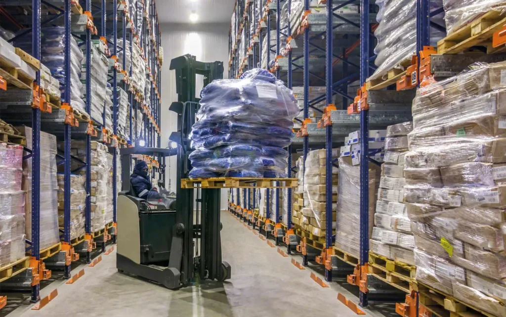 Worker operating forklift in cold storage warehouse stacking temperature-controlled pallets on tall racking