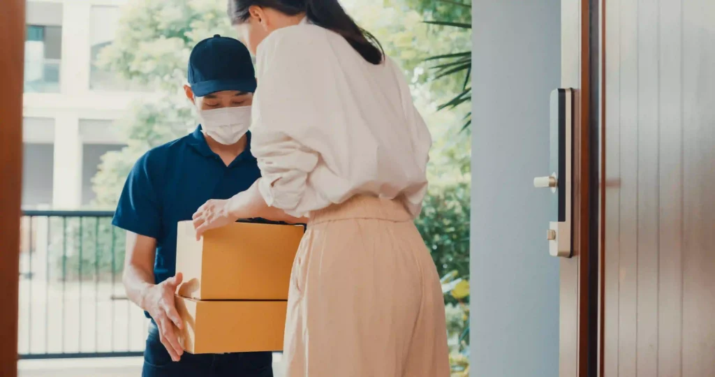 Masked courier handing two stacked cardboard boxes to a customer at her front door during a package drop off.