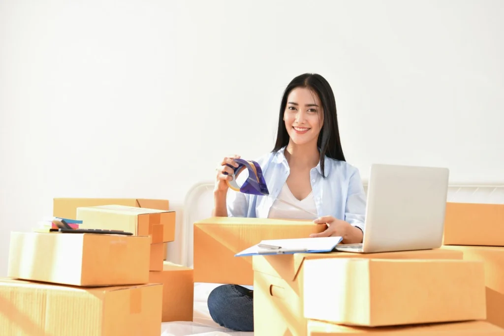 A female entrepreneur packing courier boxes with a tape dispenser and managing orders on a laptop for her delivery startup.