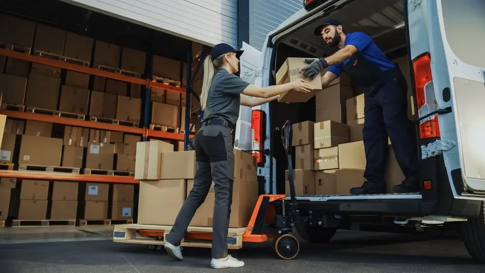 Two logistics professionals working together to load packages into a delivery van outside a warehouse, illustrating a key operational step in how to start a courier business.